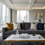 Gray sofa with four pillows in mustard, abstract navy lines on white, beige, and dark blue, in front of floor-to-ceiling windows with city views and a marble-top gold-framed coffee table