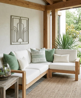 L-shaped wooden frame sofa with white cushions and green patterned and solid pillows, set on a woven rug under a wooden pergola with plants nearby and a metal wall sculpture.