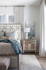 Bedroom featuring a beige headboard with circular pattern, marine spotted velvet pillow covers, a mirrored nightstand with lamp, and light beige curtains framing the window.