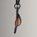 Side view of Sacronyx Necklace pendant showing a curved, light beige leaf-like form with dark edges, hanging from a black cord and ring against a gray background.