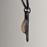 Side view of a pendant featuring a textured, semi-oval bronze element paired with a matte black, slightly curved backing, hanging from a black cord.