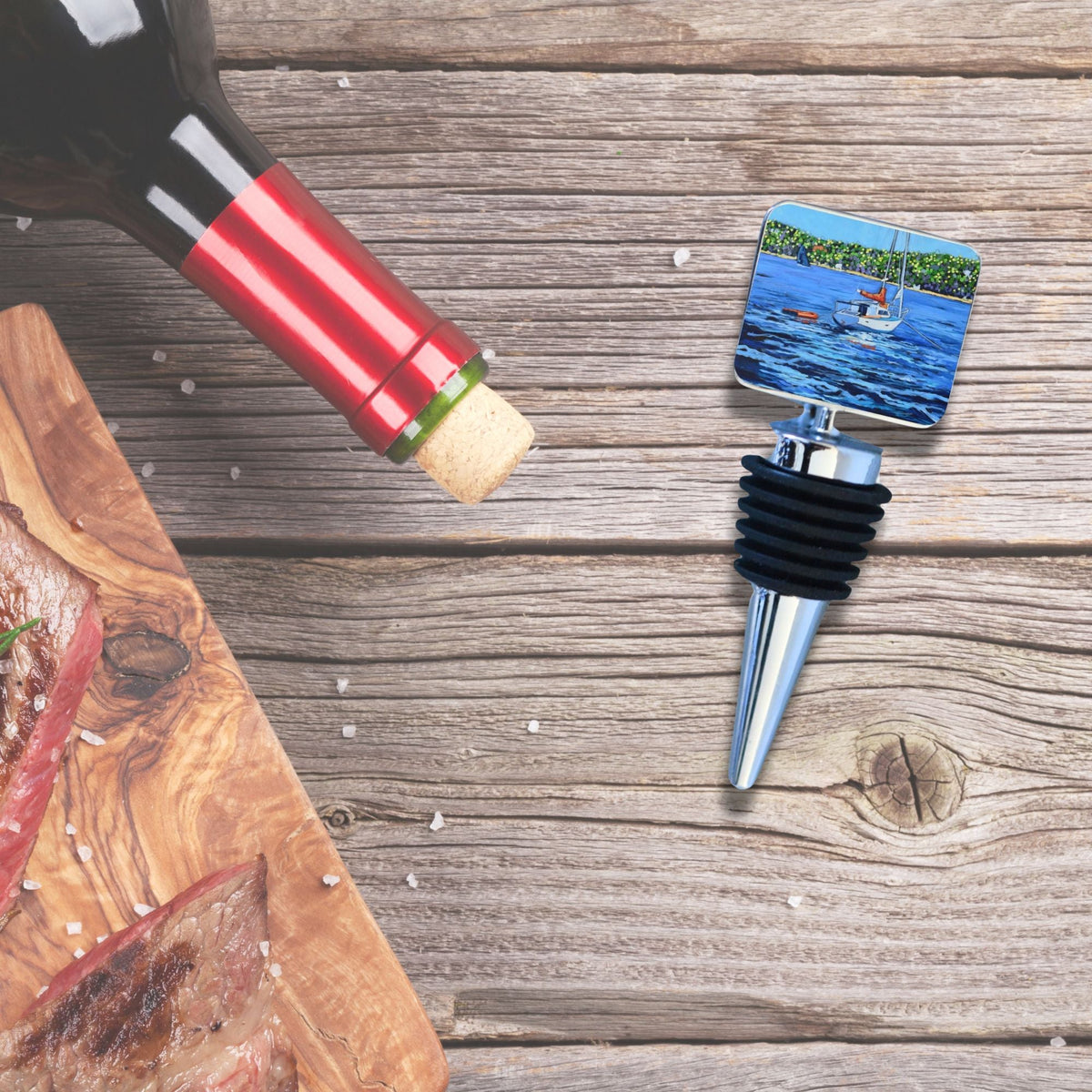 Metal wine bottle stopper with black rubber rings and a square top featuring a sailboat on blue water and green trees, sitting on a weathered wood surface beside a red wine bottle and wooden cutting board.