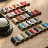 Rows of small ceramic sconces in various colors and textures including speckled, glossy, and matte finishes, displayed on a light wood surface near a green ceramic lamp base.