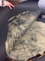 Hand lifting black sheet with gold wood grain print revealing natural beige wood cross-section with growth rings underneath, showcasing contrast between metallic and organic textures.