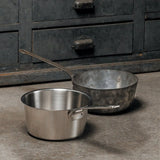 Two saucepans without lids on a concrete surface, one polished stainless steel and one with a worn, dark patina, in front of a metal cabinet with drawers.