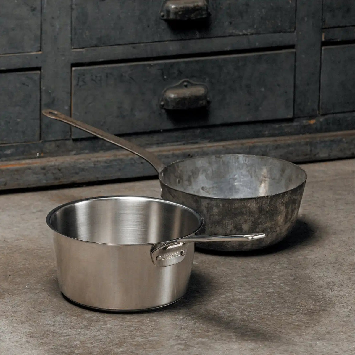 Two saucepans without lids on a concrete surface, one polished stainless steel and one with a worn, dark patina, in front of a metal cabinet with drawers.