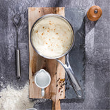 Top view of a saucepan with creamy sauce sprinkled with pepper, resting on a two-tone wood cutting board beside a small white pitcher, a whisk, peppercorns, and flour on a gray textured surface.