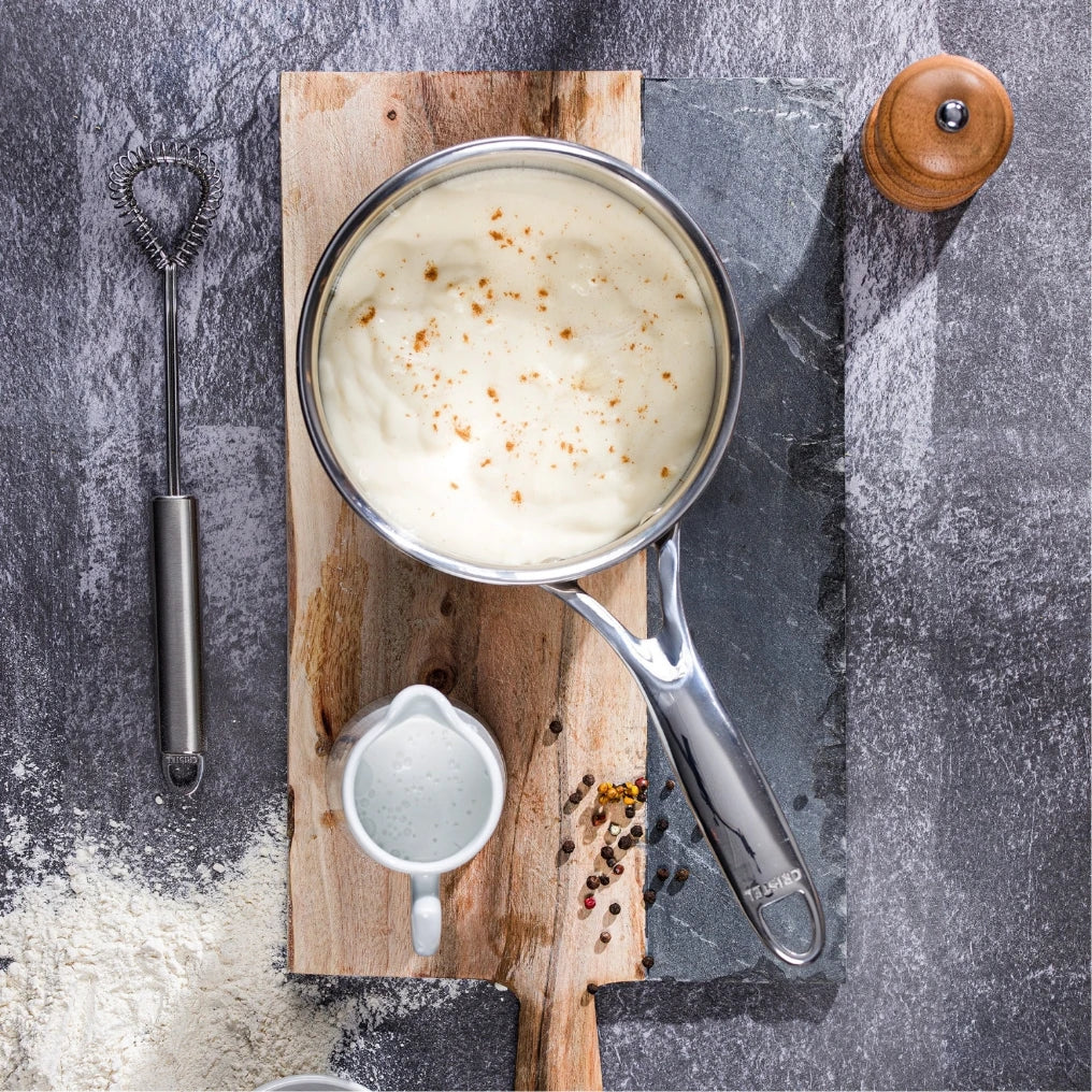 Top view of a saucepan with creamy sauce sprinkled with pepper, resting on a two-tone wood cutting board beside a small white pitcher, a whisk, peppercorns, and flour on a gray textured surface.