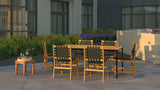 Outdoor dining set with wooden table and six armless chairs featuring black woven webbing seats and backs, arranged on a spacious patio with greenery and purple flowers in the background.