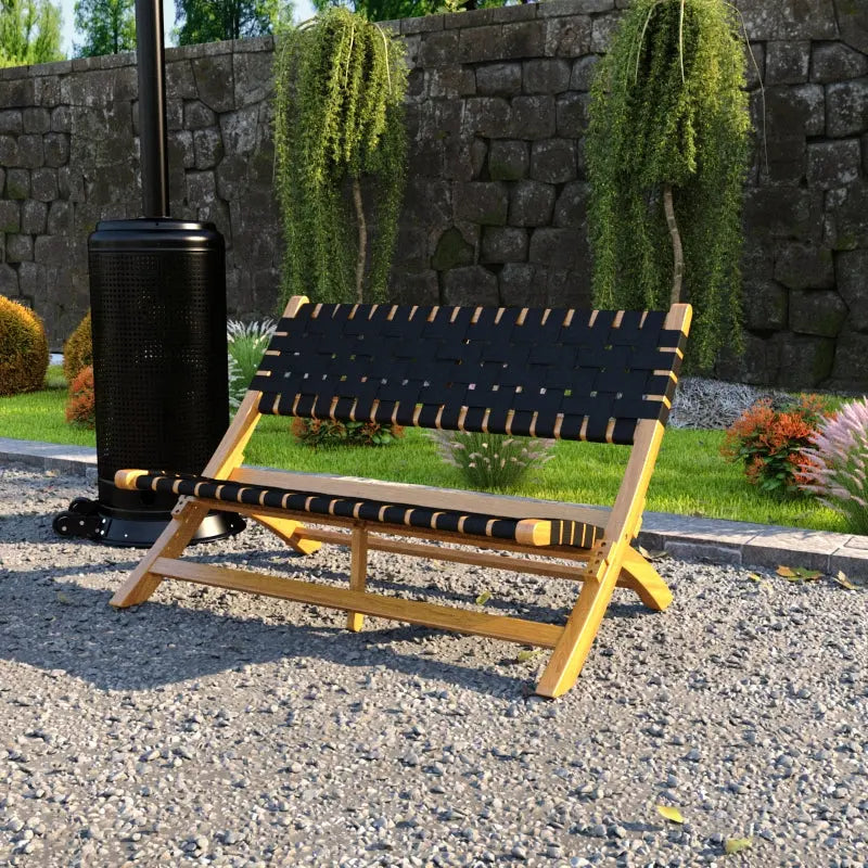 Outdoor wooden bench with black woven seat and backrest straps, set on gravel with a stone wall and greenery in the background.