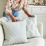 Two square pillows with celadon paisley and geometric patterns on a white couch, a person arranging one pillow while wearing a floral blouse and jeans.