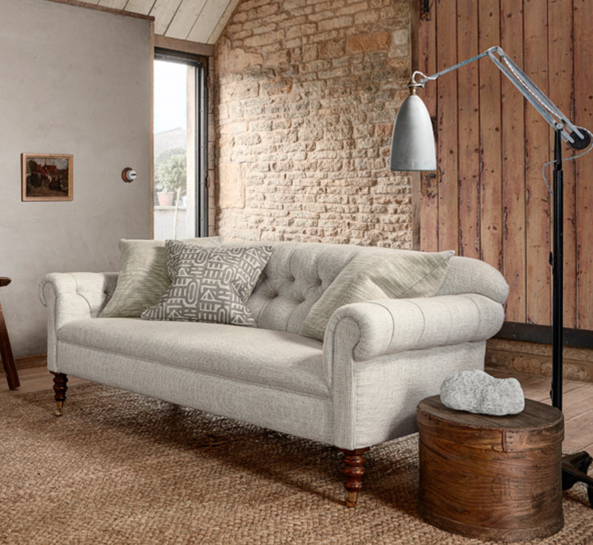 Light grey upholstered sofa with rolled arms and three patterned cushions, set against a textured stone wall and large wooden door, with a metal floor lamp nearby.