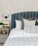 Bed with a vertically paneled blue upholstered headboard, white bedding, and three decorative pillows in grey and patterned fabrics against a wallpapered wall with grey leaf motifs.