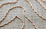 Close-up of oyster-colored fabric with shiny golden thread embroidery forming a wavy, abstract pattern over a textured woven background.