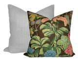 Square pillow with a brown background featuring colorful floral and leaf patterns in green, blue, red, and yellow, positioned in front of a plain light gray pillow.