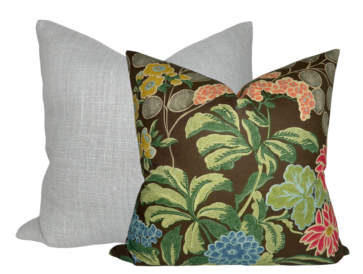 Square pillow with a brown background featuring colorful floral and leaf patterns in green, blue, red, and yellow, positioned in front of a plain light gray pillow.