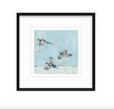 Fine art print of three pheasants in mid-flight against a soft blue sky with subtle cream tones, framed in a simple black frame with white matting.
