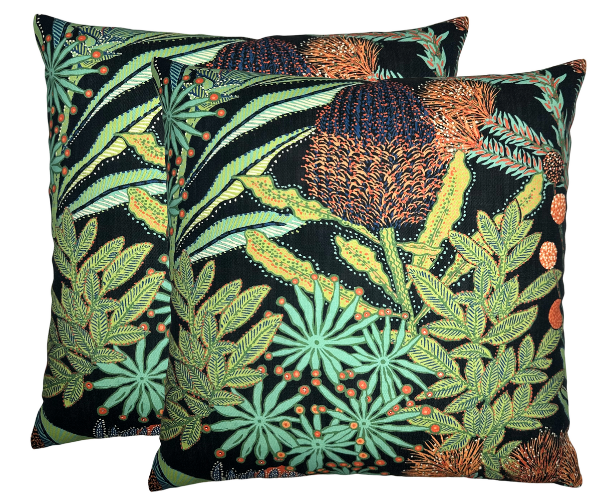 Two square black pillow covers featuring detailed botanical patterns with orange protea flowers and green leafy foliage in varied shades against a dark background.