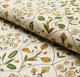 Close-up of flax-colored fabric with a repeating pattern of stylized floral motifs in green, mustard, and blue, outlined in black on a light natural background.