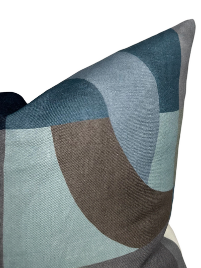 Close-up of a geometric pillow cover featuring overlapping curved shapes in indigo, muted blue, gray, and brown tones on textured fabric.