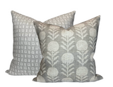 Two square pillows with sand grey fabric, one featuring a repetitive white geometric pattern and the other with a white stylized floral design.