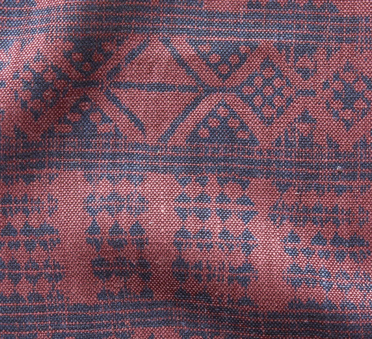 Close-up of woven fabric with intricate geometric pattern in dark blue on a dusty rose background, showing textured threads and subtle folds.