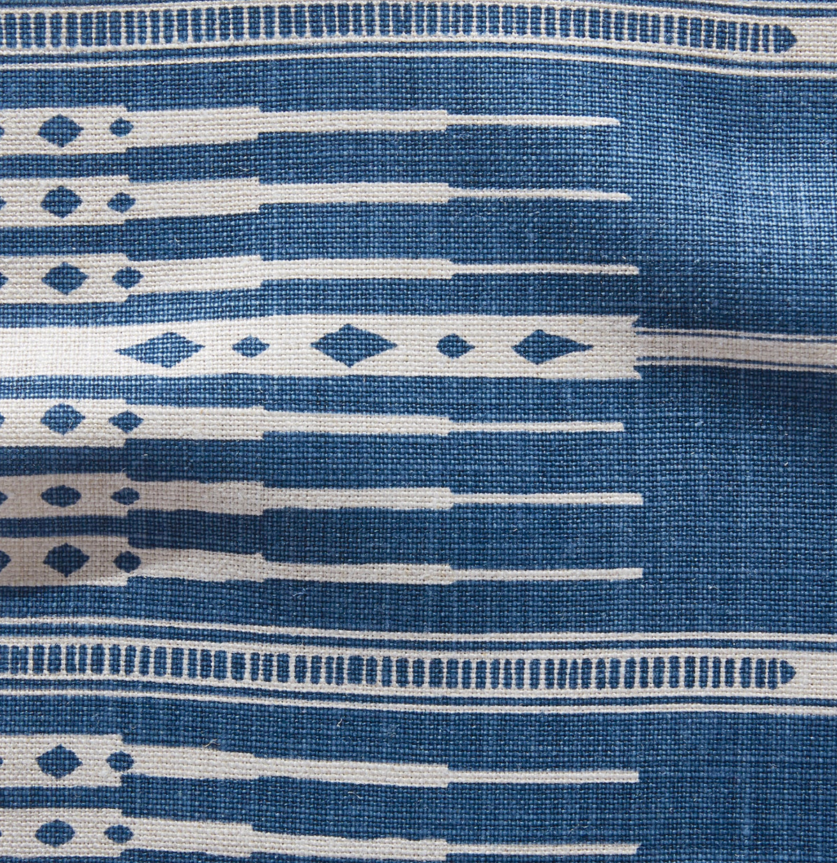 Close-up of a woven textile in indigo blue with horizontal rows of elongated white shapes and diamond motifs, showing subtle fabric texture and natural wrinkles.