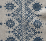 Indigo geometric and floral embroidery pattern on natural beige fabric featuring diamond shapes and circular motifs with intricate cross-stitch details.