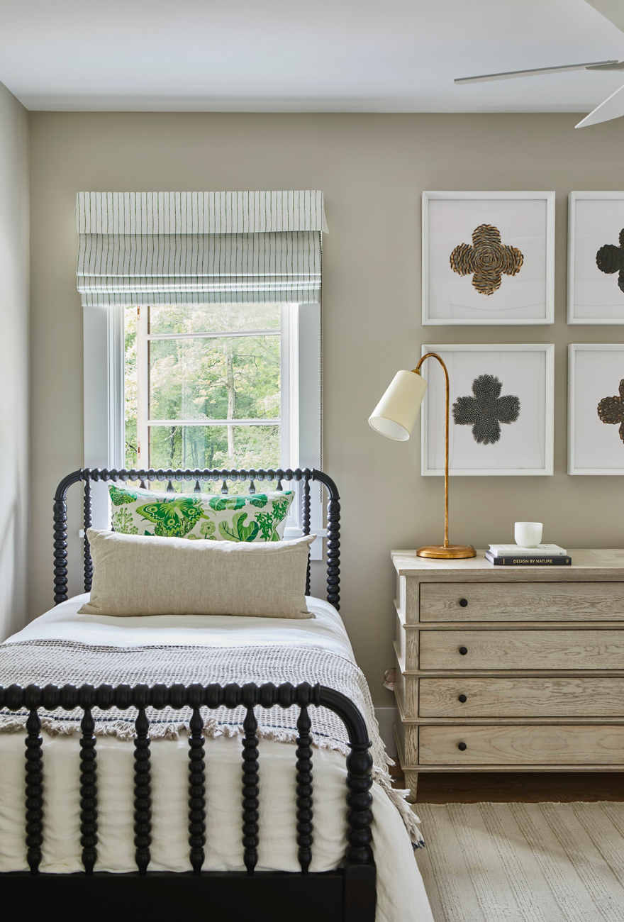 Single black spindle bed with layered pillows including a green butterfly leaf-patterned pillow, beside a light wood chest of drawers under four framed botanical prints.