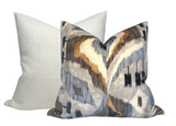 Two square pillows with one solid cream textured pillow behind a front pillow featuring a multicolored abstract pattern in gray, brown, cream, and blue tones.