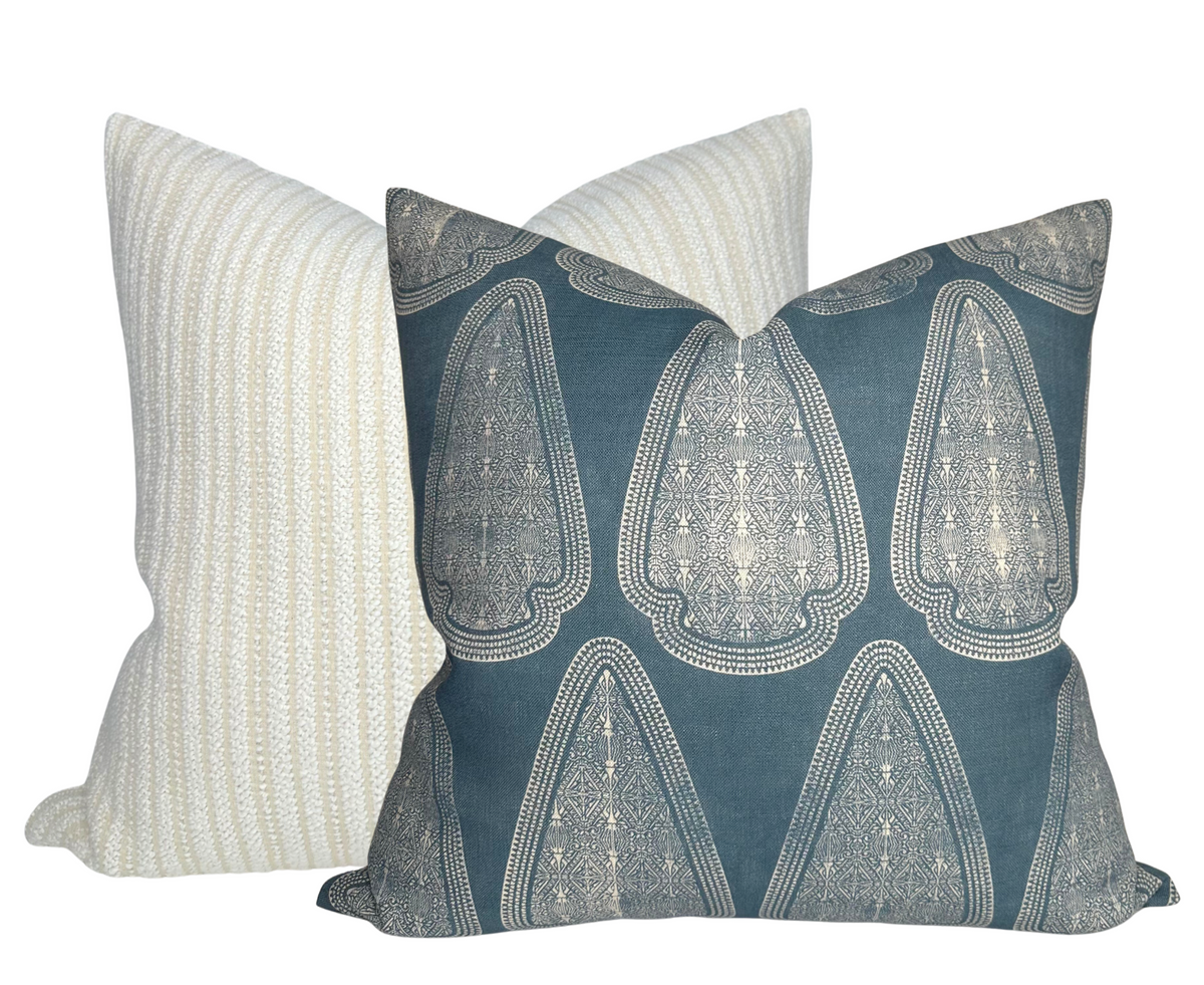 Square pillow with a teal background featuring symmetrical, intricate cream lace-like patterns, placed in front of a beige pillow with vertical textured stripes.