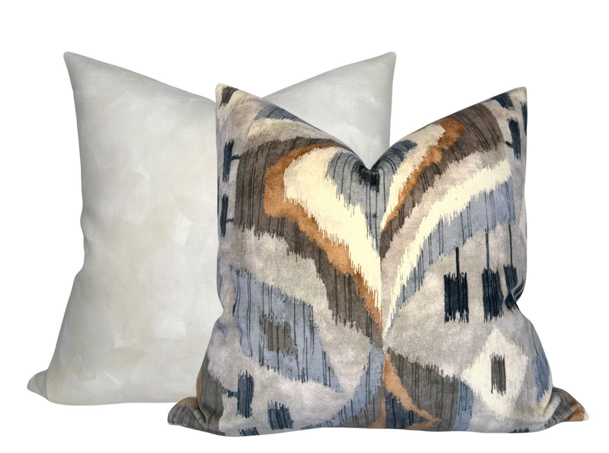Two square pillow covers shown, one with a soft pearl white solid color and the other with a flowing abstract pattern in muted blues, grays, browns, and creams.
