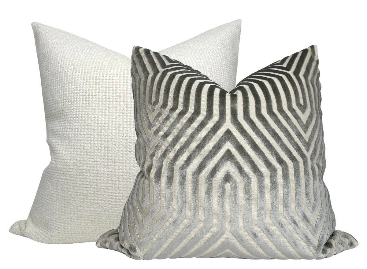 Two square pillows with knife edges; front pillow has a grey and white geometric chevron pattern, the back pillow is solid pale grey with a textured weave.