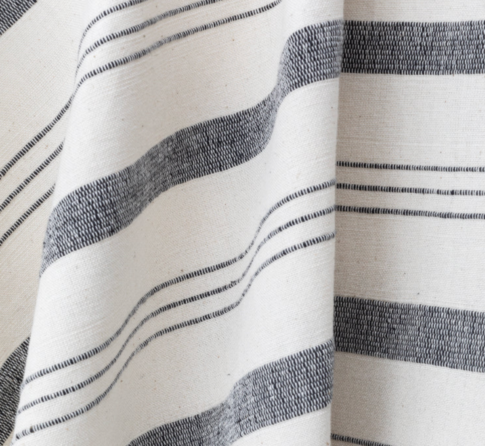 Natural-colored fabric with alternating thick and thin black horizontal stripes, showing woven texture and soft draping folds.
