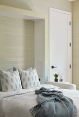 Bed with white sheets and pillows featuring a blue geometric Myriad pattern, complemented by a draped lake blue throw blanket in a softly lit bedroom corner.