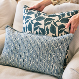 Two decorative pillows on a beige fabric chair, one with a blue and cream abstract leaf pattern being held, the other with a blue geometric eye-shaped pattern resting on the seat.