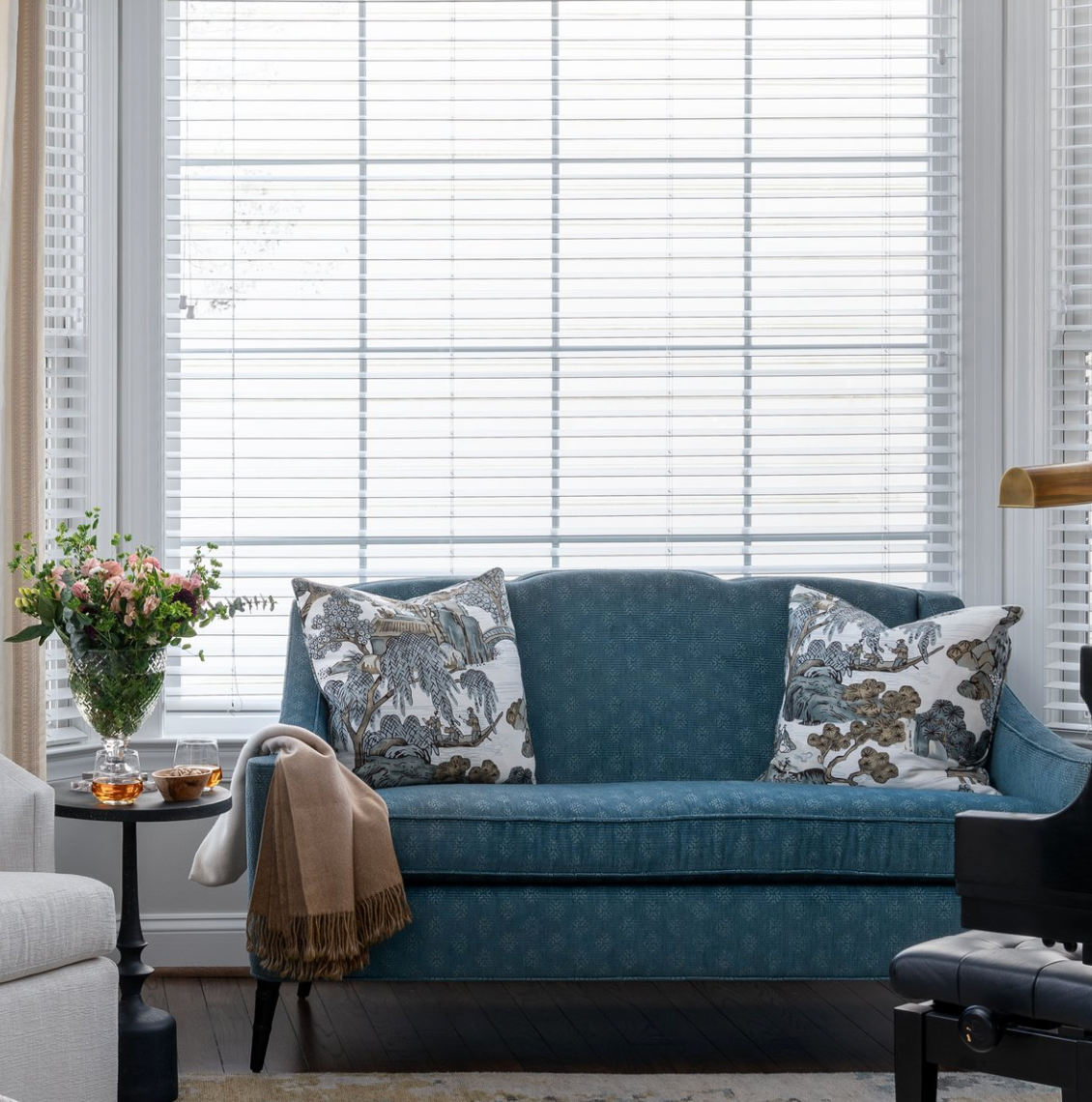 Blue patterned loveseat with two grey Asian scenic patterned pillows, a beige throw over one arm, beside a small black side table with flowers in a vase, in front of a window with white blinds.