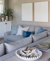 Gray sectional sofa with patterned blue, white, and solid blue pillows arranged against textured beige wall with two empty wooden frames above.