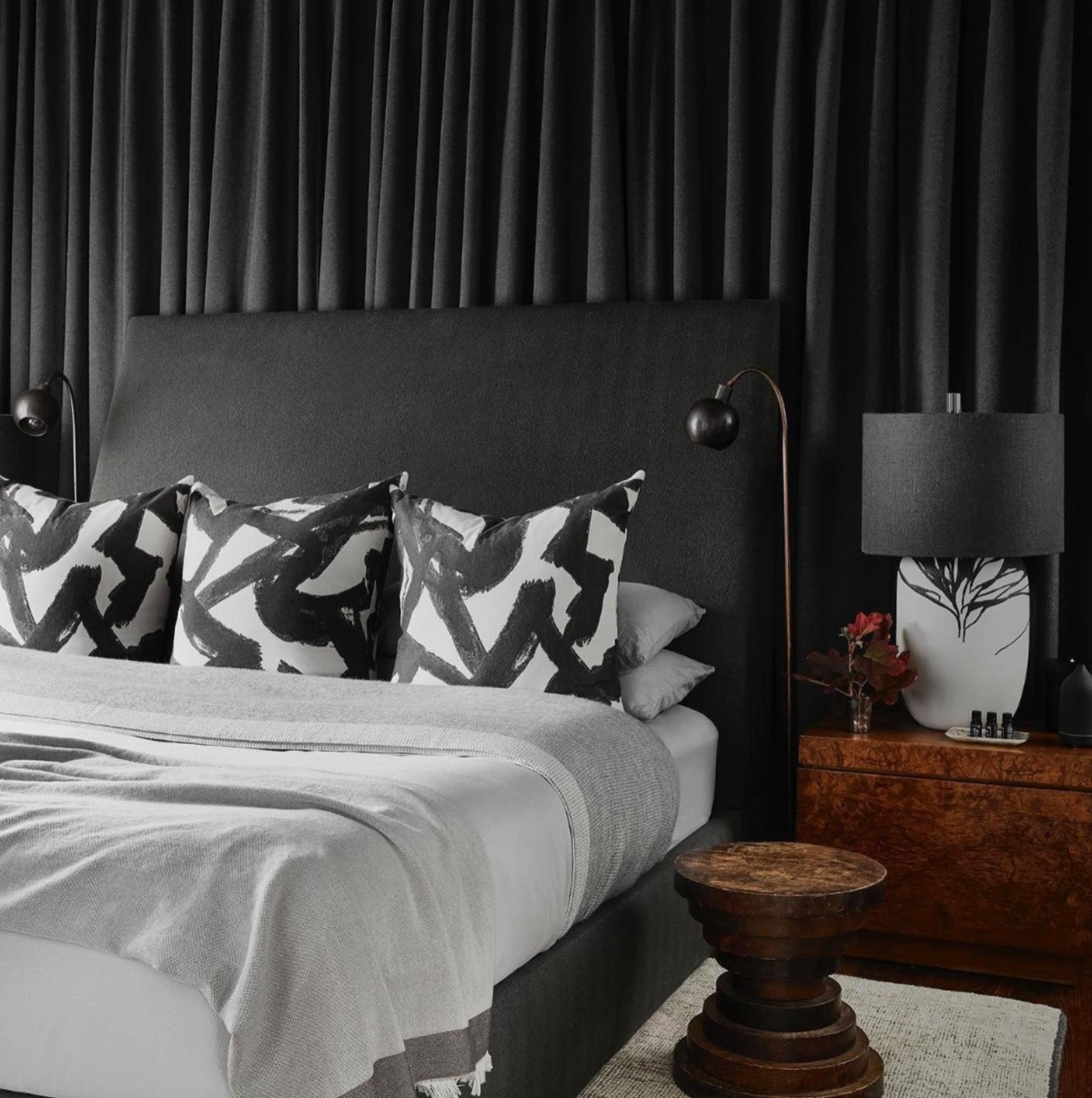 King-size bed with dark gray upholstered headboard, three black and white abstract brushstroke pillows, gray bedding, flanked by dark lamps and a wooden nightstand with a white vase and black tree design.