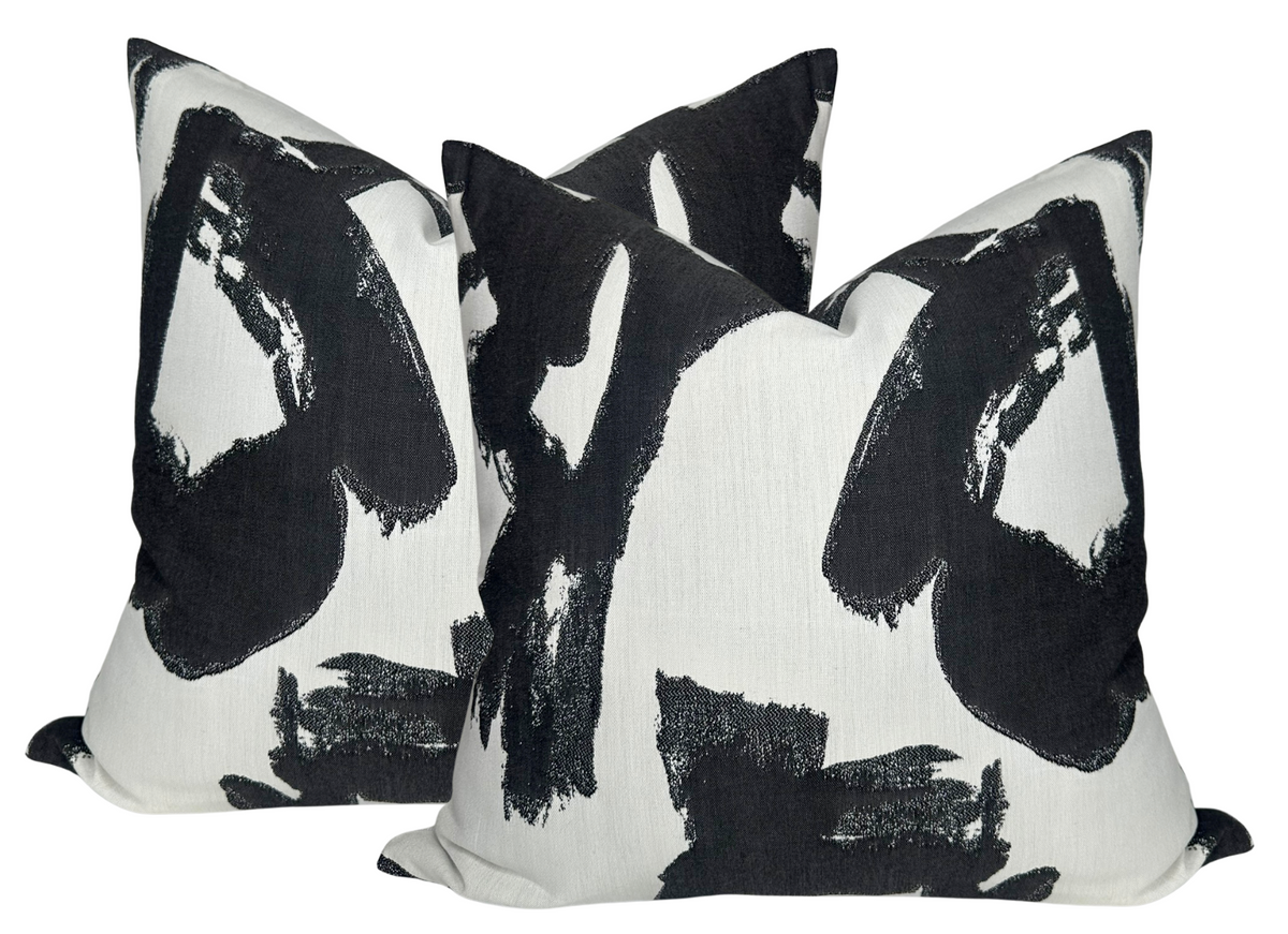 Two square pillow covers with an abstract black brushstroke pattern on a white background, arranged overlapping and viewed from the front.