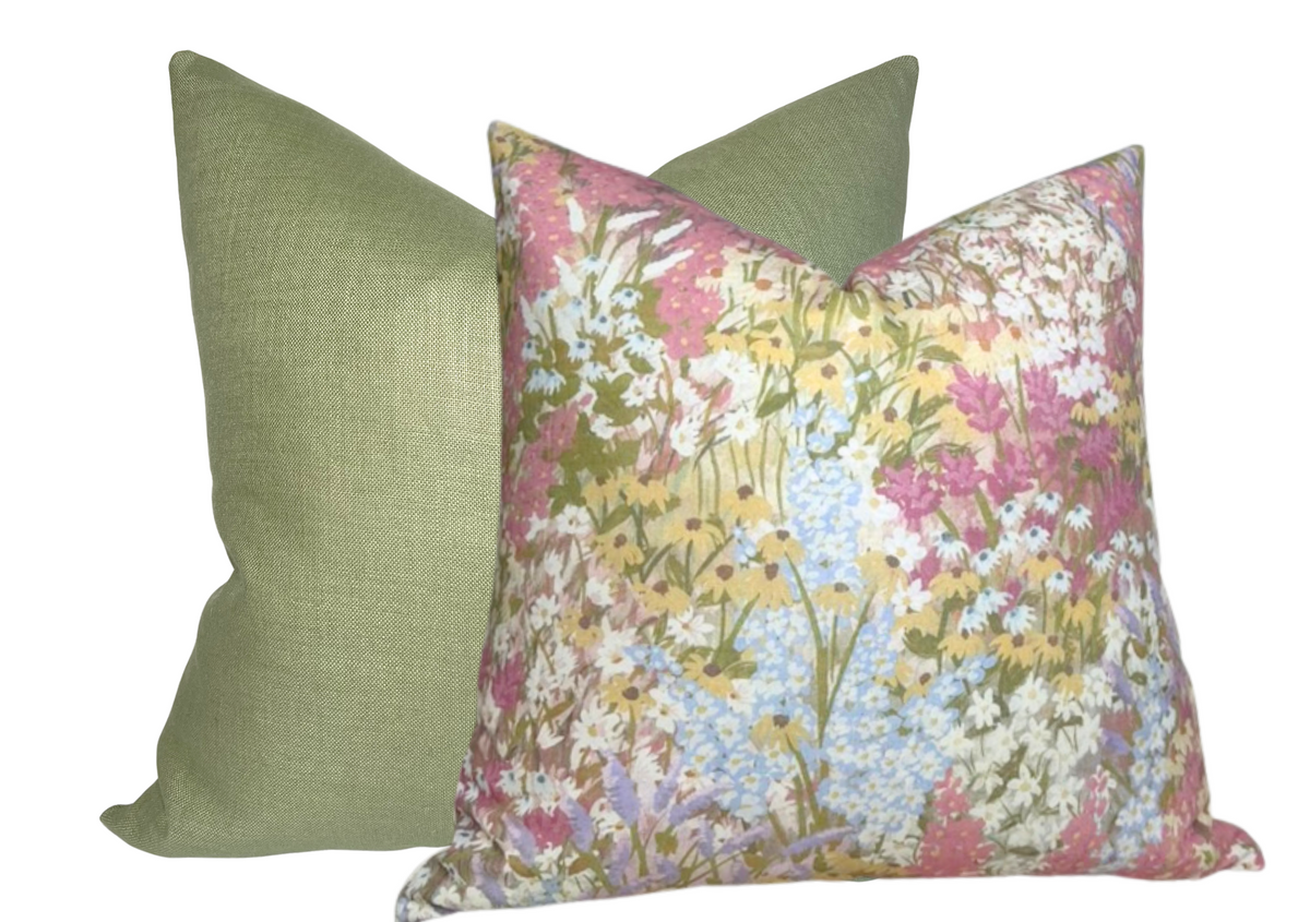 Square pillow with a floral pattern in blush tones and pastel colors, positioned in front of a solid muted green square pillow.