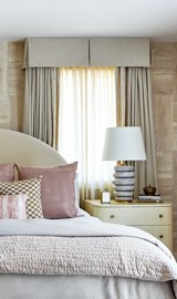 Bedroom window with beige curtains and valance, flanked by a side table holding a ribbed gray lamp with white shade and books, beside a bed with mauve and patterned pillows.