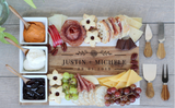 Rectangular wooden charcuterie board engraved with names and date, topped with cheeses, grapes, crackers, meats, chocolate, and floral-shaped cookies, accompanied by three wooden-handled cheese knives.