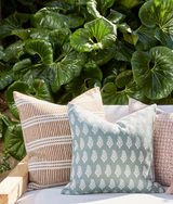 Three decorative pillows with geometric and leaf patterns in soft blue, beige, and muted red, arranged on a light wooden outdoor bench against large green leaves.