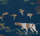 Pillow fabric with a white and brown hunting dog pointing amidst grass and plants on a navy blue background with flying reddish-brown and white game birds.