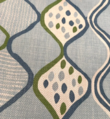 Close-up of blue fabric with a wavy geometric pattern featuring diamond shapes outlined in green and blue, filled with multi-colored small ovals in blue, green, and gray tones.
