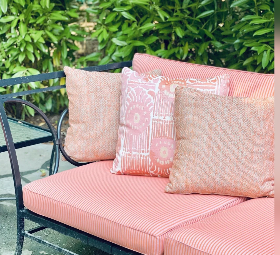 Outdoor bench with coral and white striped cushions and three decorative pillows in mixed melon coral patterns, set against green leafy background.