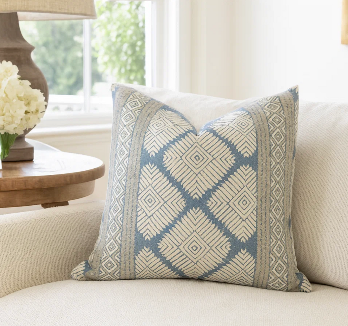 Square pillow with blue and beige geometric diamond patterns resting on a cream fabric armchair near a wooden side table with a lamp and white flowers.