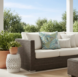 Outdoor seating with dark woven rattan sofa featuring cream cushions, accented by a blue pillow with a botanical print, surrounded by potted green plants on a patio.