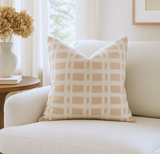 Square pillow with a soft pink grid pattern on a light cream background, placed on a white upholstered sofa beside a wooden side table with a white vase and dried flowers.